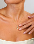 Woman wearing a gold necklace with a pendant, hand touching her chest.