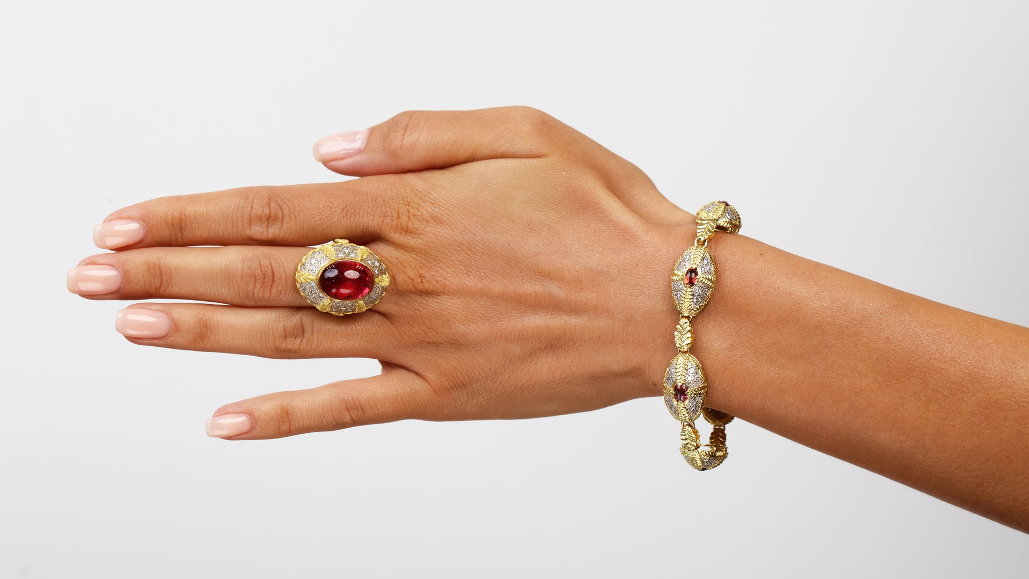 Hand wearing a gold ring and bracelet with red gemstones on a light gray background