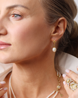 Close-up of a blond woman wearing pearl earrings and a necklace against a neutral background.